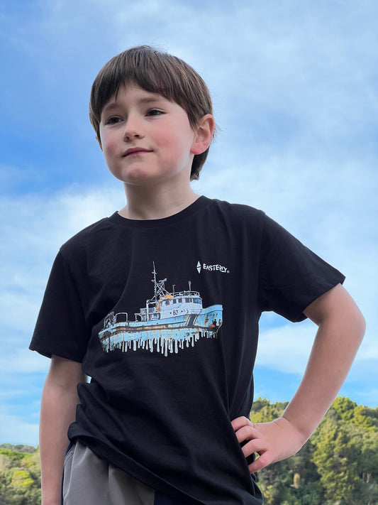 A young boy standing outdoors against a blue sky, wearing a black EASTERLY kids' tee with a fishing boat illustration on the front and a small white EASTERLY chest logo