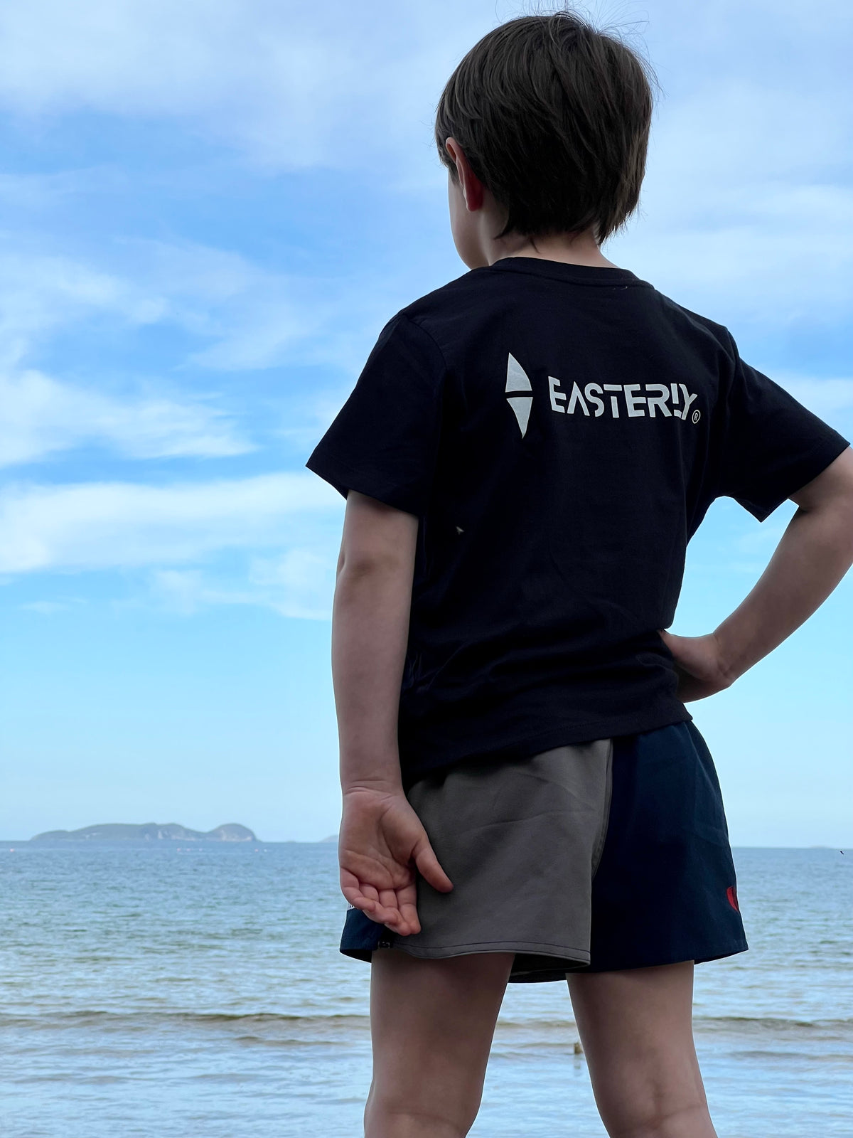 A child standing at the water's edge on a Stewart Island beach, looking out to sea, wearing a black EASTERLY kids' tee showing the white EASTERLY icon and wordmark on the back