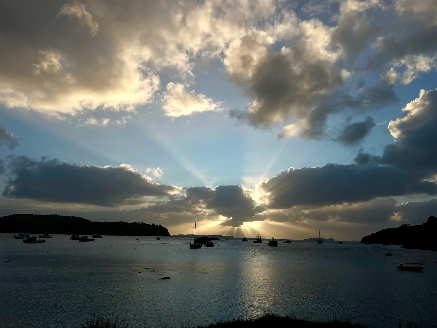 Light rays breaking through storm clouds over Halfmoon Bay, Rakiura — Stewart Island, New Zealand. Boats silhouetted on the calm water at dusk, islands in the distance.