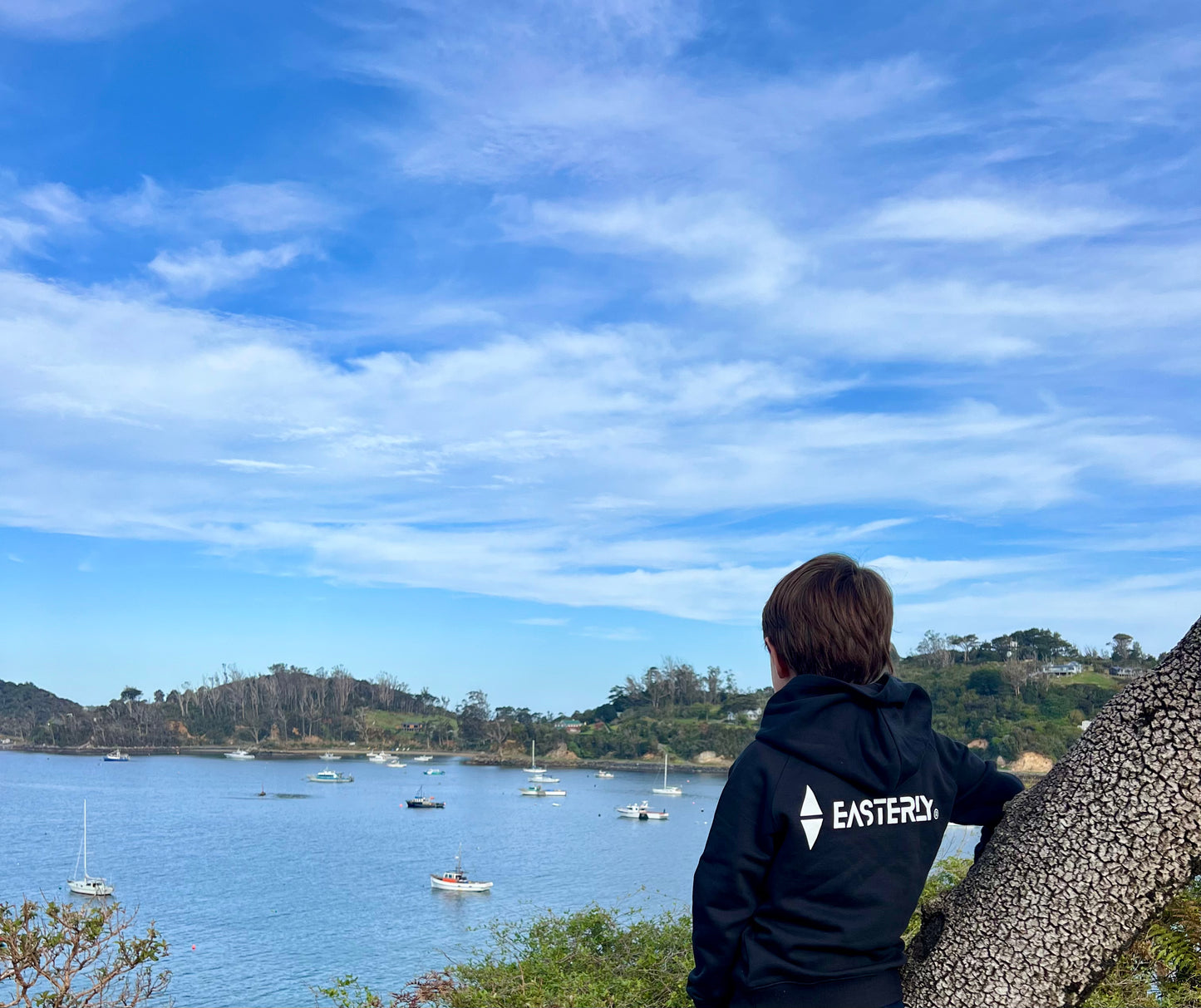 Veaux wearing a black Sammy Seal kids’ hoodie with the white EASTERLY back logo, looking East in an East wind across the bay dotted with boats.