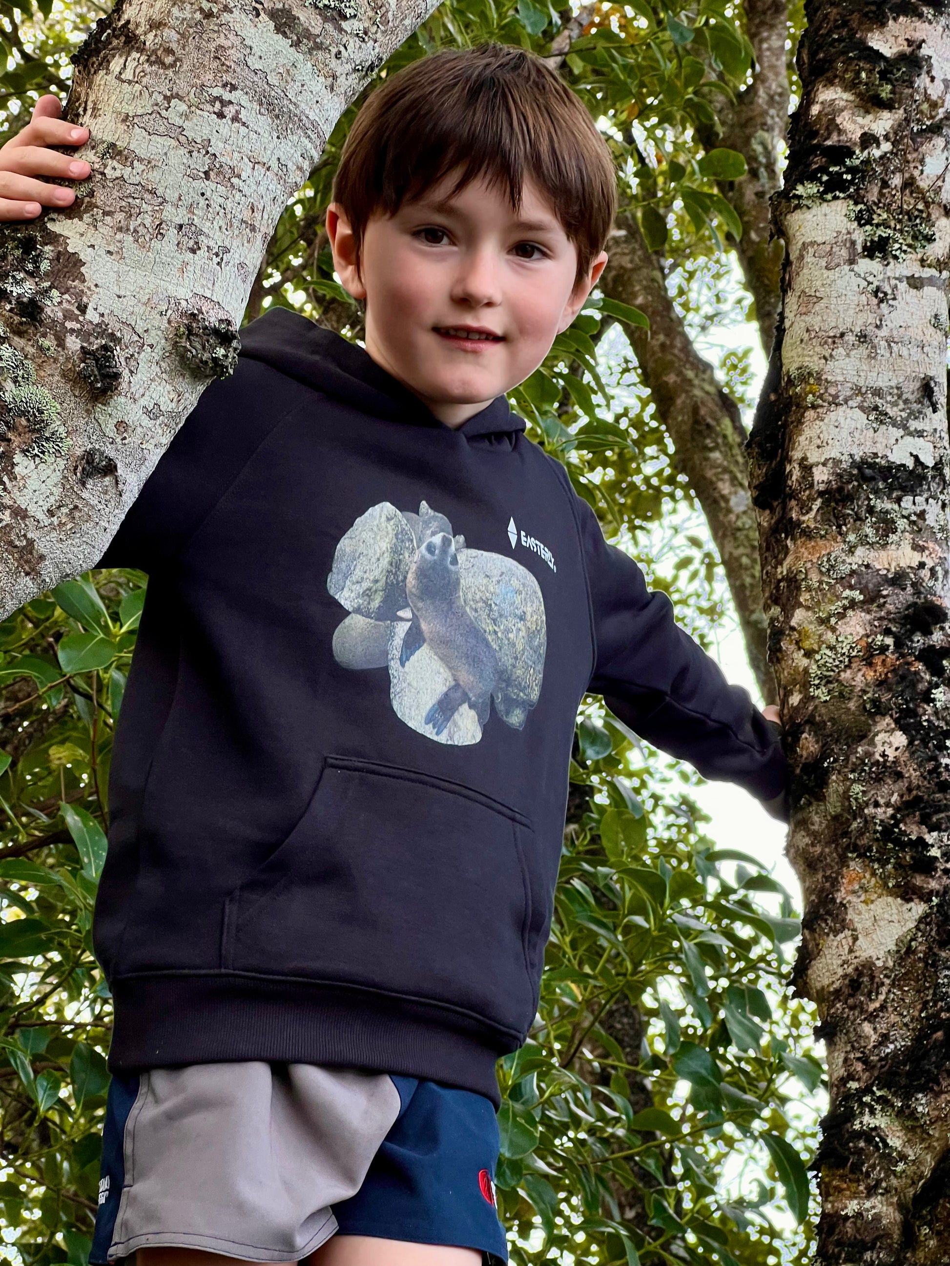 Veaux standing between tree trunks in a black Sammy Seal kids’ hoodie, showing the front print and pouch pocket.