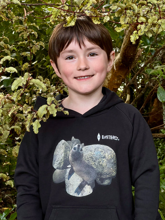 Veaux smiling among leafy branches, wearing a black Sammy Seal kids’ hoodie with the seal pup print.