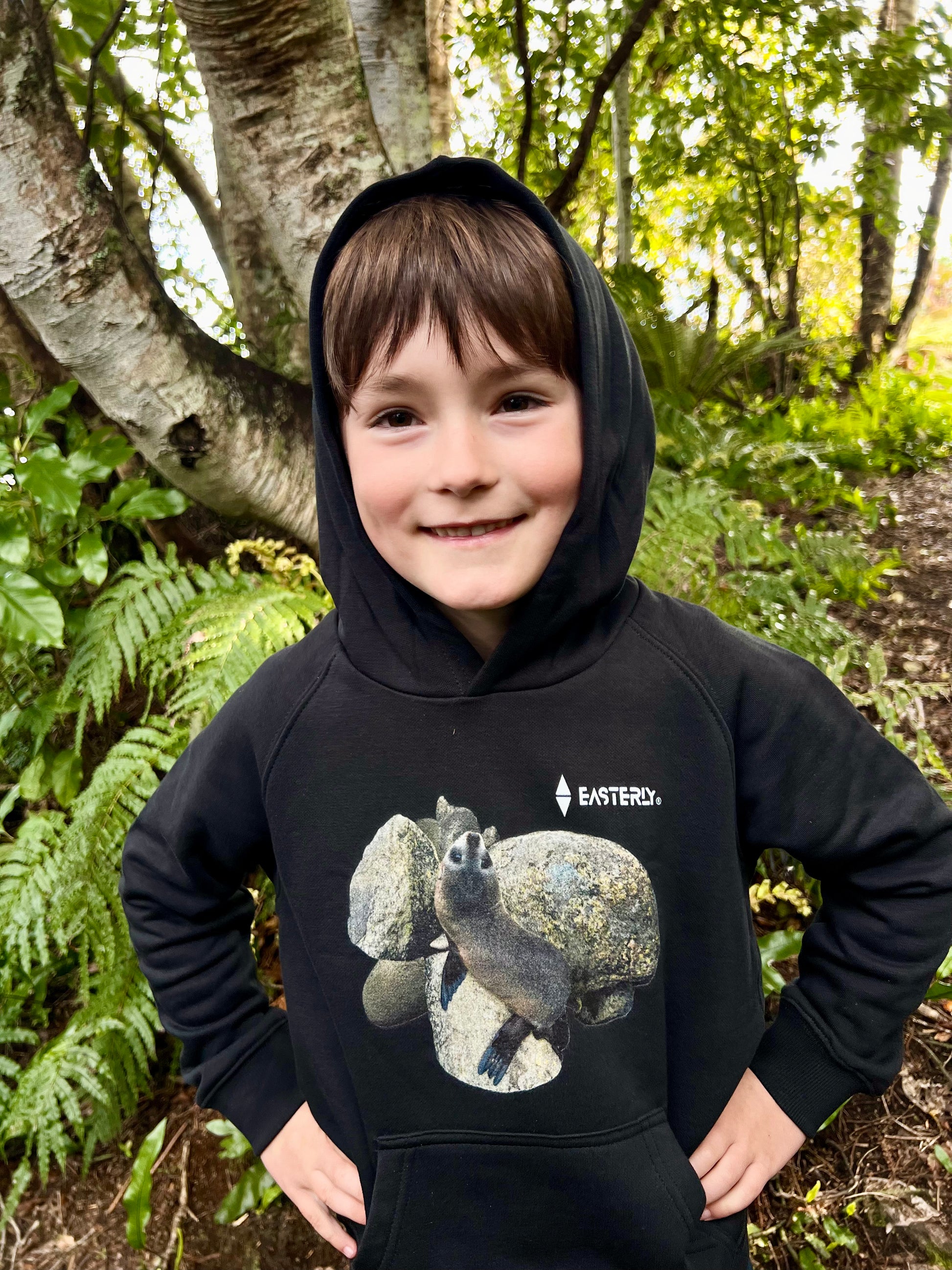 Veaux wearing a black EASTERLY Sammy Seal kids’ hoodie among trees and ferns.