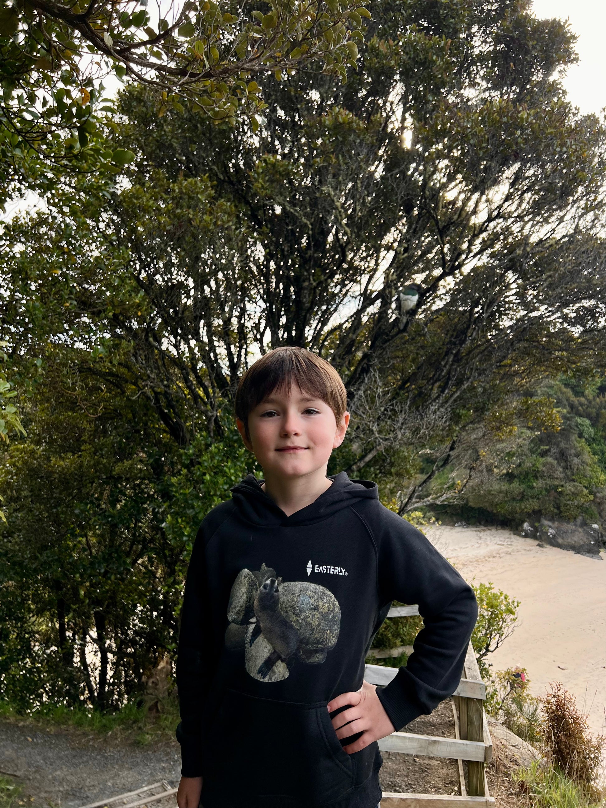 Veaux wearing a black Sammy Seal kids’ hoodie beside wooden steps overlooking  Bathing Beach, Stewart Island.