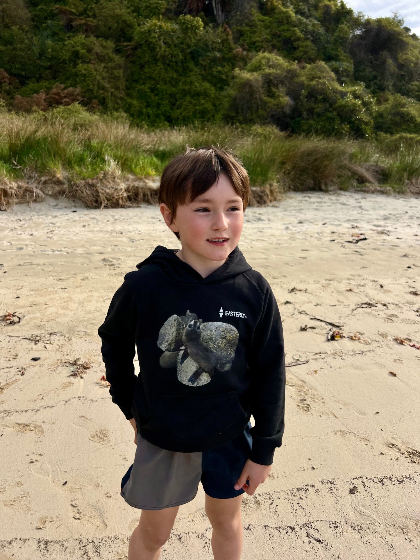 Veaux standing on sandy Bathing Beach, Stewart Island in a black Sammy Seal kids’ hoodie with a seal pup and rocks print.
