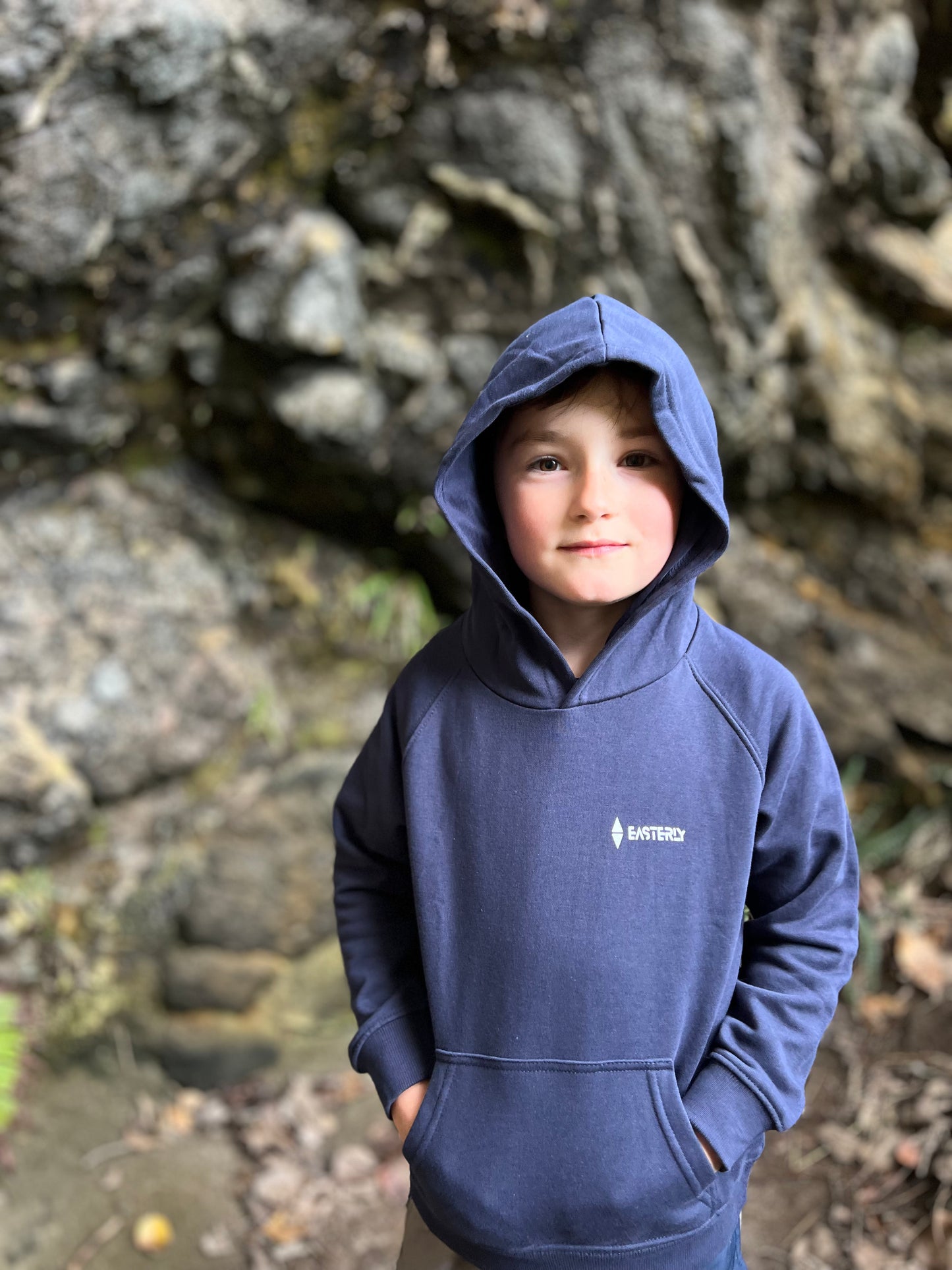 A young boy wearing a navy blue hooded sweatshirt with a small white Easterly logo on the left chest, standing outdoors in front of a rocky, blurred background. Bathing Beach, Stewart Island.