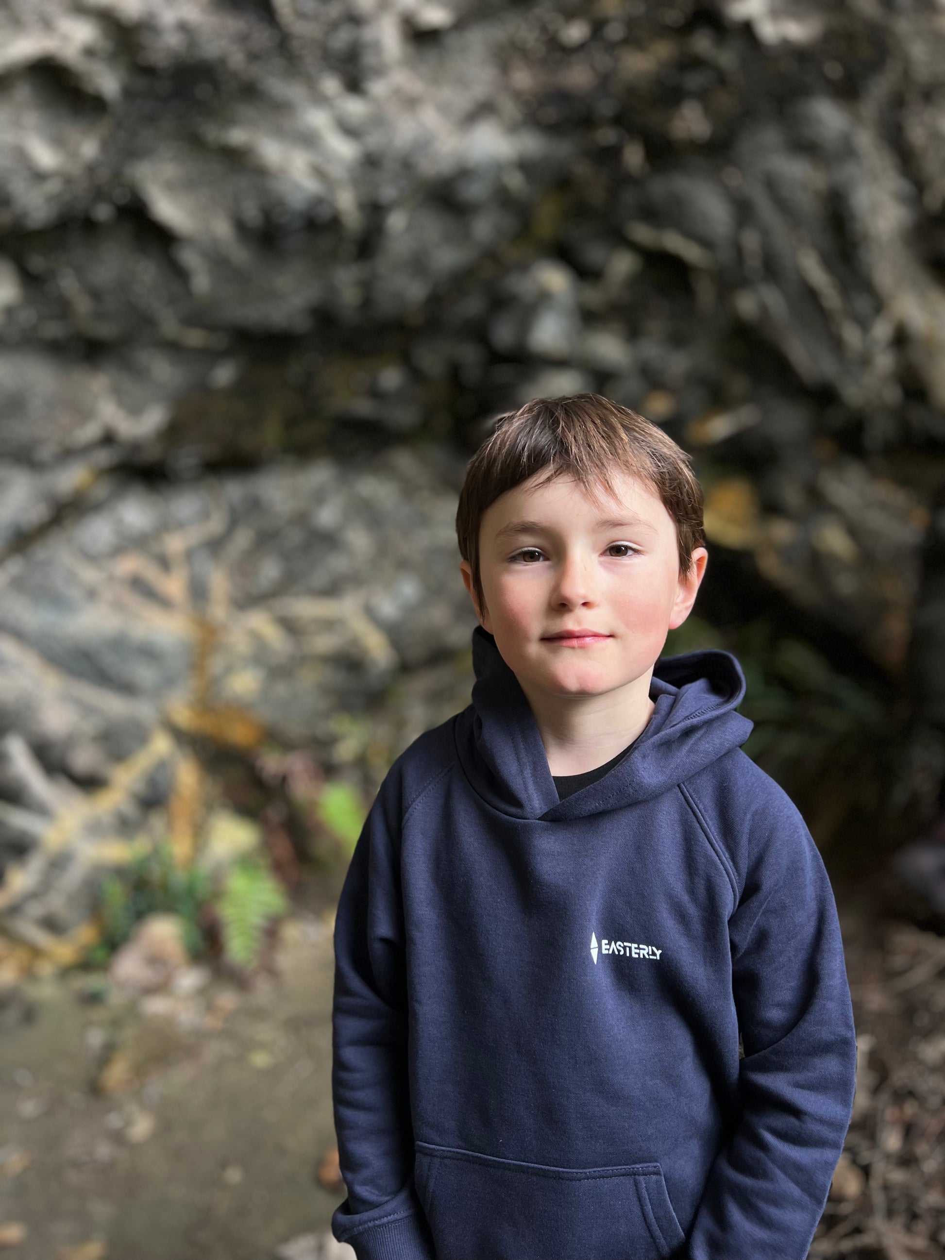 A young boy with short brown hair stands in front of a rocky outdoor background, wearing a navy blue hoodie with a small white Easterly logo on the left chest.