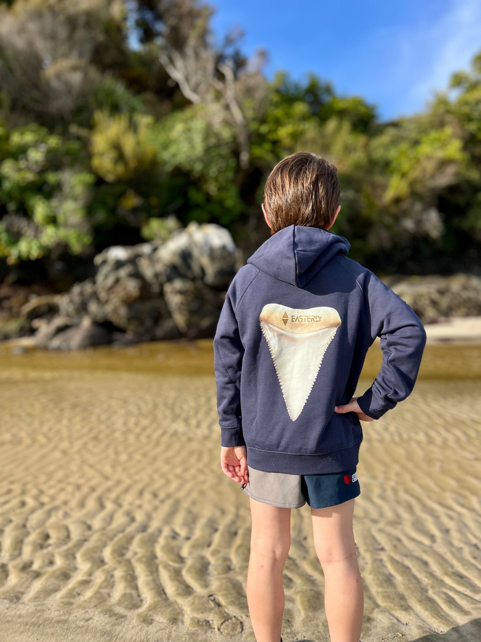A child standing on a sandy beach facing a rocky shoreline, wearing a navy blue hoodie with a large shark tooth graphic on the back. Bathing Beach Stewart Island.