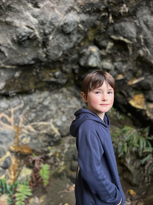 A young boy wearing a navy blue hoodie stands in front of a rocky outdoor cave wall with green ferns visible in the background.