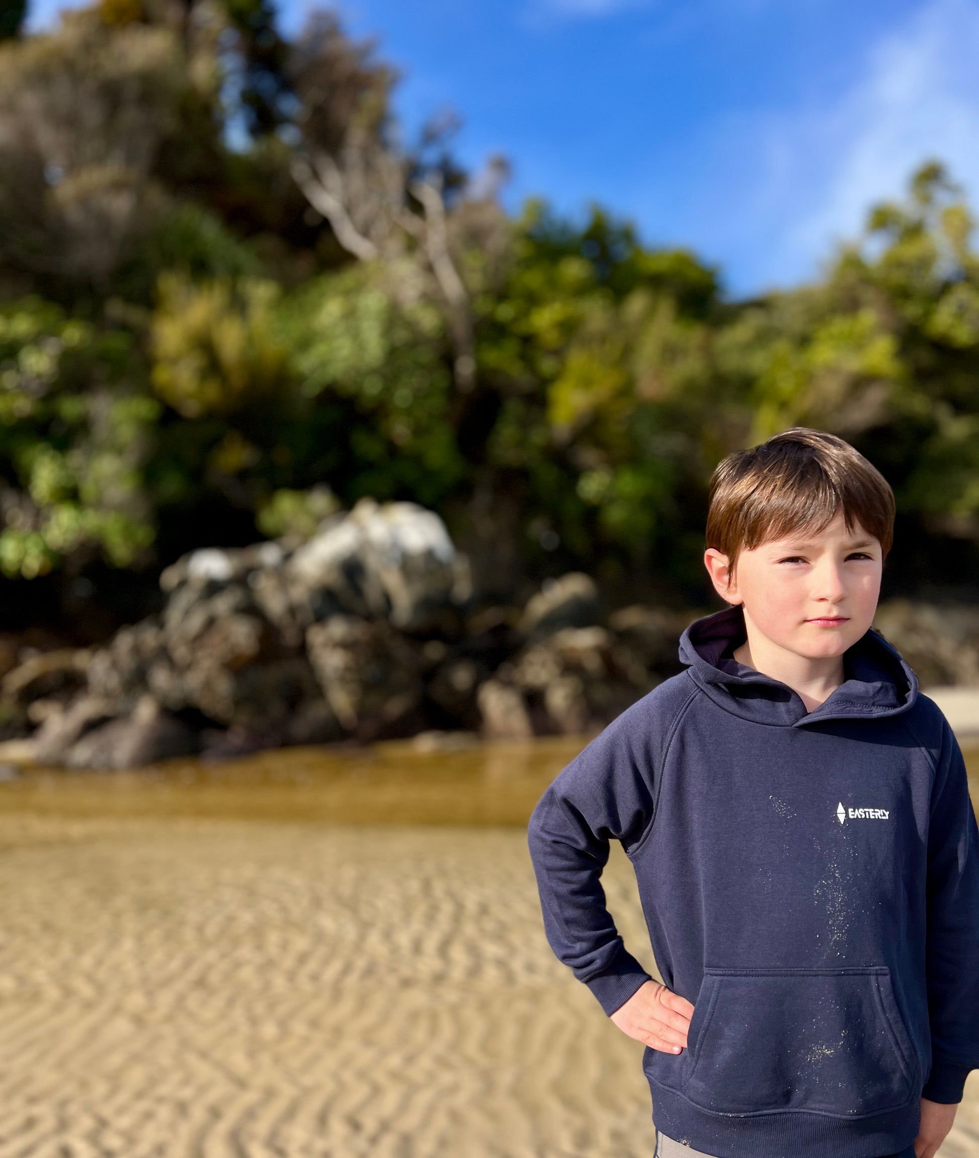 A young boy wearing a navy blue hoodie with a small white Easterly logo stands on Bathing Beach Stewart Island, with rocks and native trees in the background.