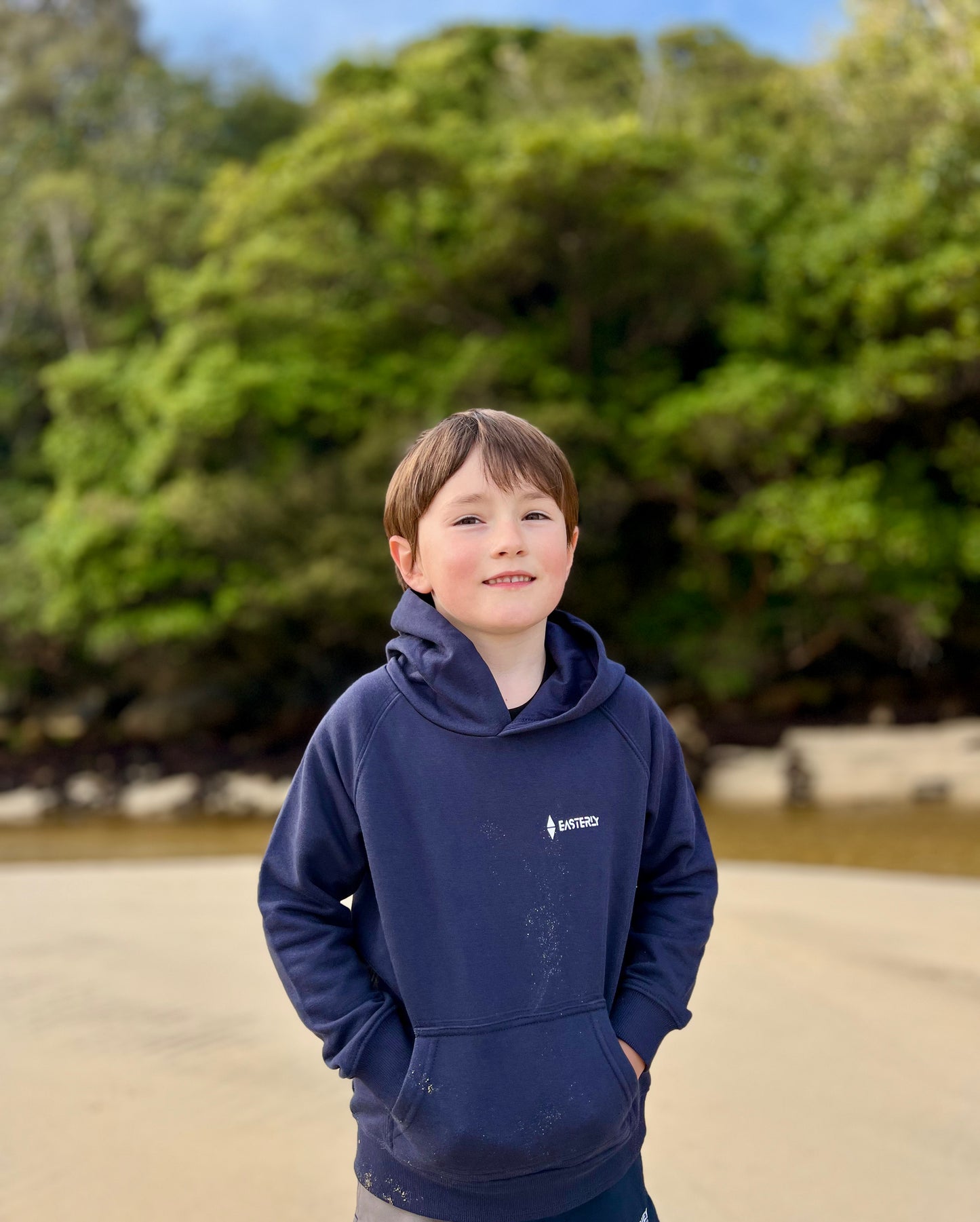 A young boy wearing a navy blue Easterly hoodie stands outdoors with a blurred background of green trees and a sandy ground. Bathing Beach , Stewart Island.
